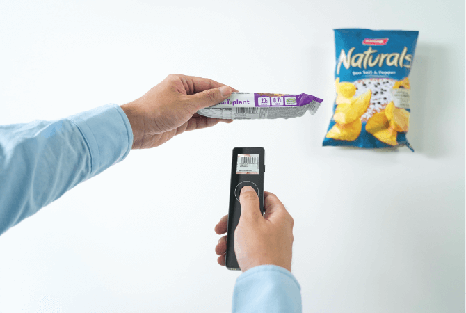 a hand holds the Elys Eye Touch, an Elys accessory, with their right hand and scans a barcode of a granola bar with their left hand. The barcode is reflected onto the Elys Eye screen. The background is a white table and there is a bag of Naturals potato chips on the table in the upper-right corner.