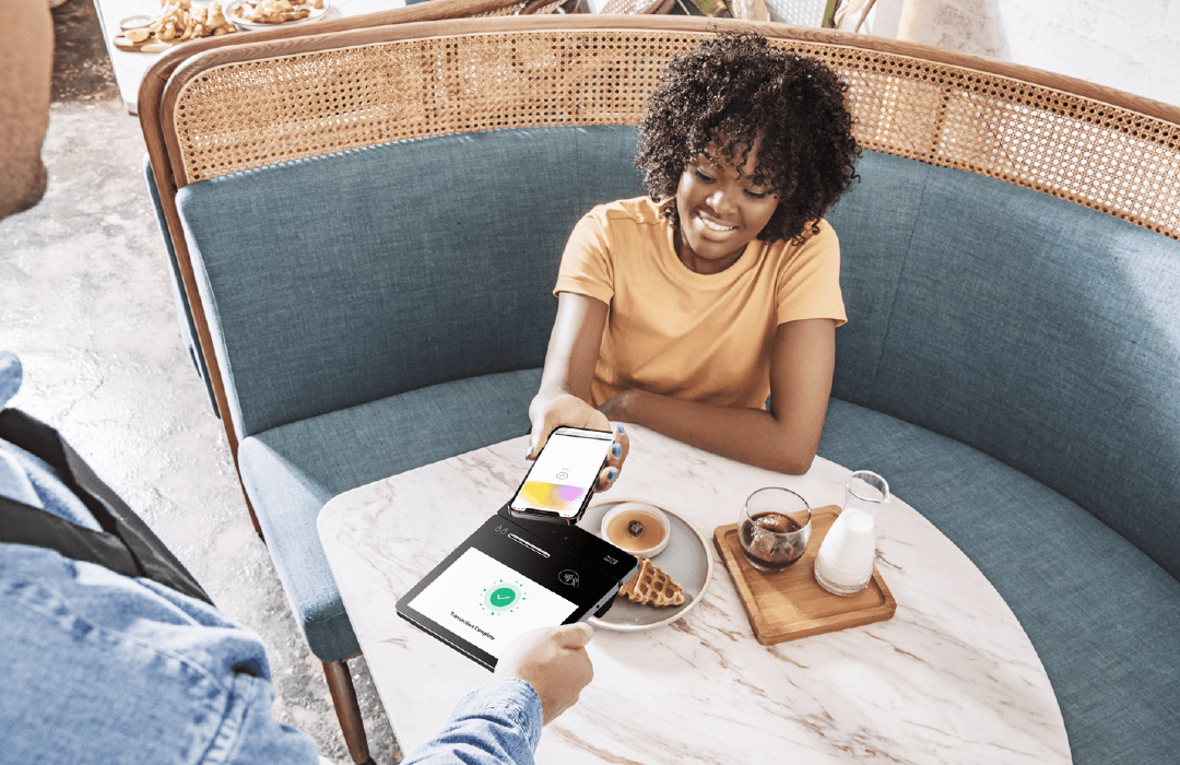 A women wearing a yellow shirt who is sitting in a teal colored booth is paying for her meal on her phone by tapping the Elys tablet, a mobile payment device.