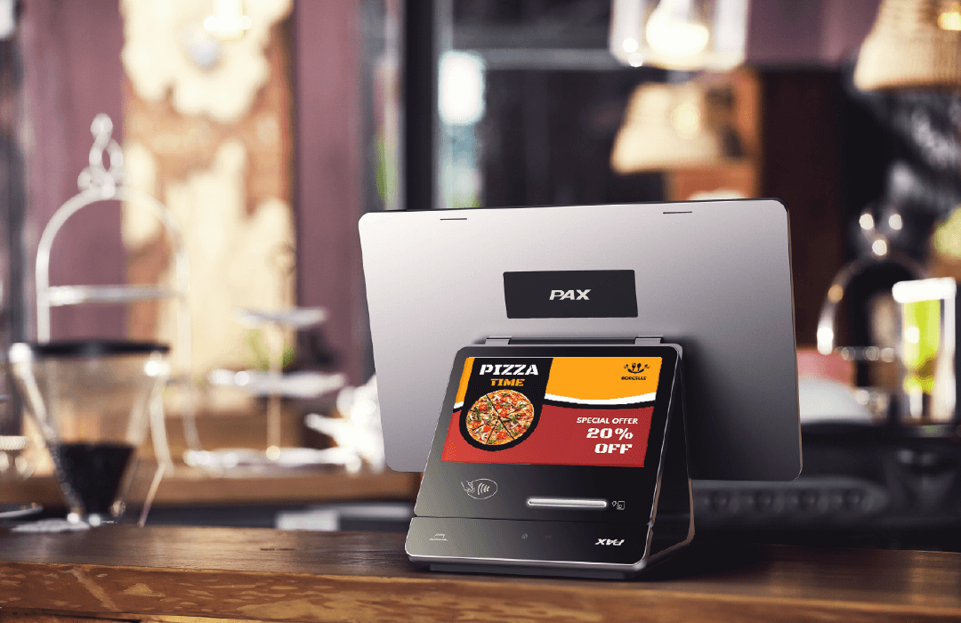 The Elys Workstation with the Elys Tablet, a payment device, sits on a wooden counter of a pizza shop. The Elys Tablet displays an ad for the pizza shop offering 20% off.